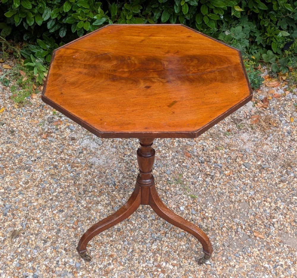 An antique Regency mahogany occasional table featuring an octagonal tilt-top surface, a turned central pedestal stem, three splayed tripod legs, and brass castors. View II