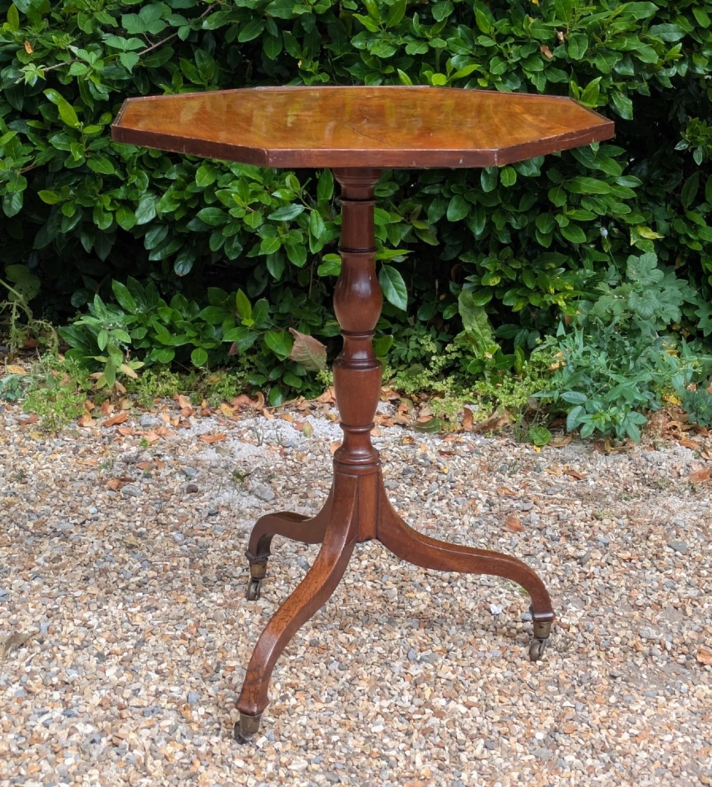 An antique Regency mahogany occasional table featuring an octagonal tilt-top surface, a turned central pedestal stem, three splayed tripod legs, and brass castors. View V