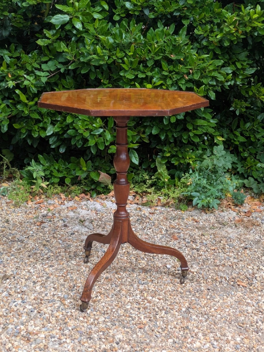 An antique Regency mahogany occasional table featuring an octagonal tilt-top surface, a turned central pedestal stem, three splayed tripod legs, and brass castors. View VIII
