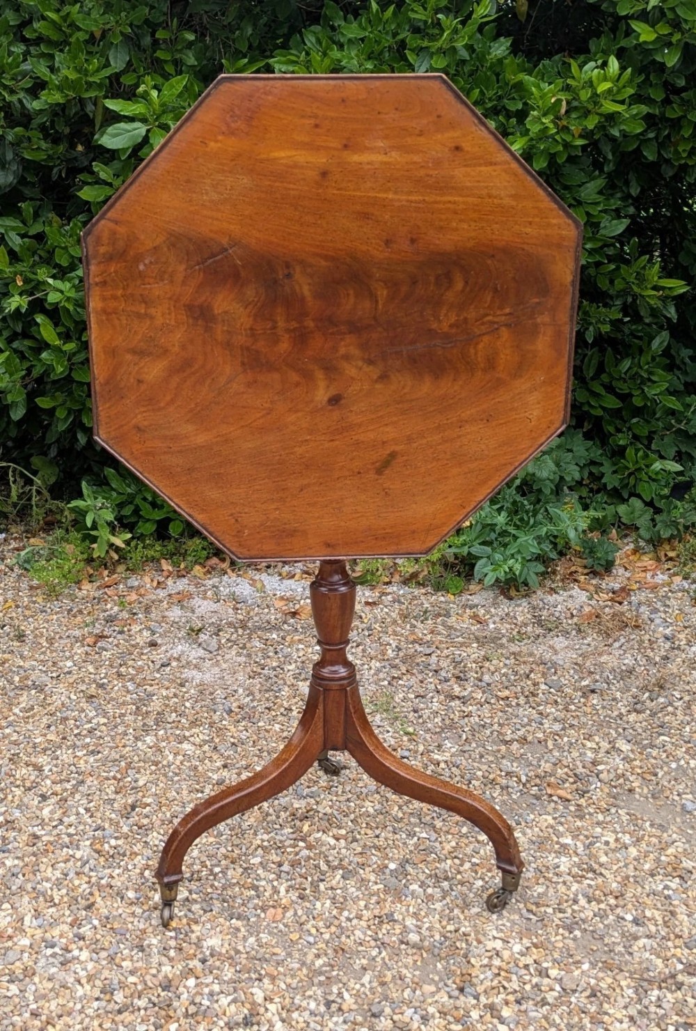 An antique Regency mahogany occasional table featuring an octagonal tilt-top surface, a turned central pedestal stem, three splayed tripod legs, and brass castors. View IX