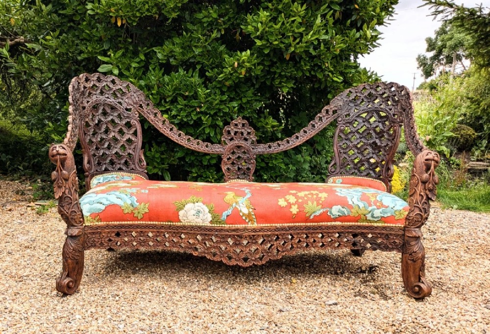 An antique Anglo-Indian carved hardwood sofa featuring an ornate openwork pierced backrest, carved lion armrests, and a patterned coral upholstered seat.