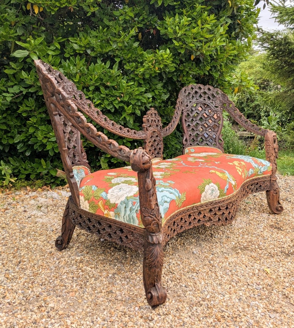 An antique Anglo-Indian carved hardwood sofa featuring an ornate openwork pierced backrest, carved lion armrests, and a patterned coral upholstered seat. View II