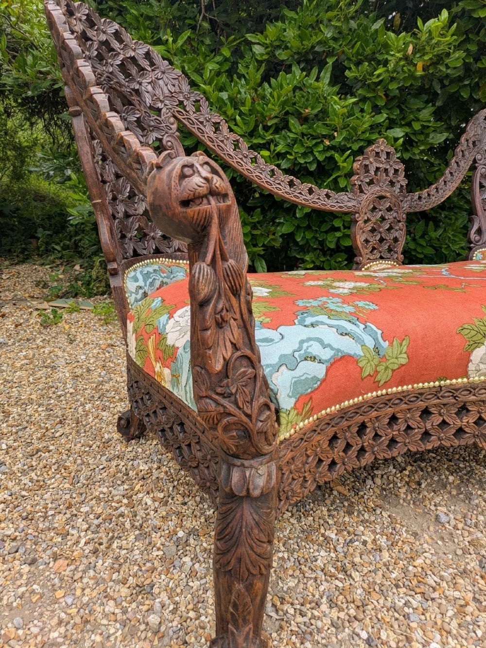 An antique Anglo-Indian carved hardwood sofa featuring an ornate openwork pierced backrest, carved lion armrests, and a patterned coral upholstered seat. View IV