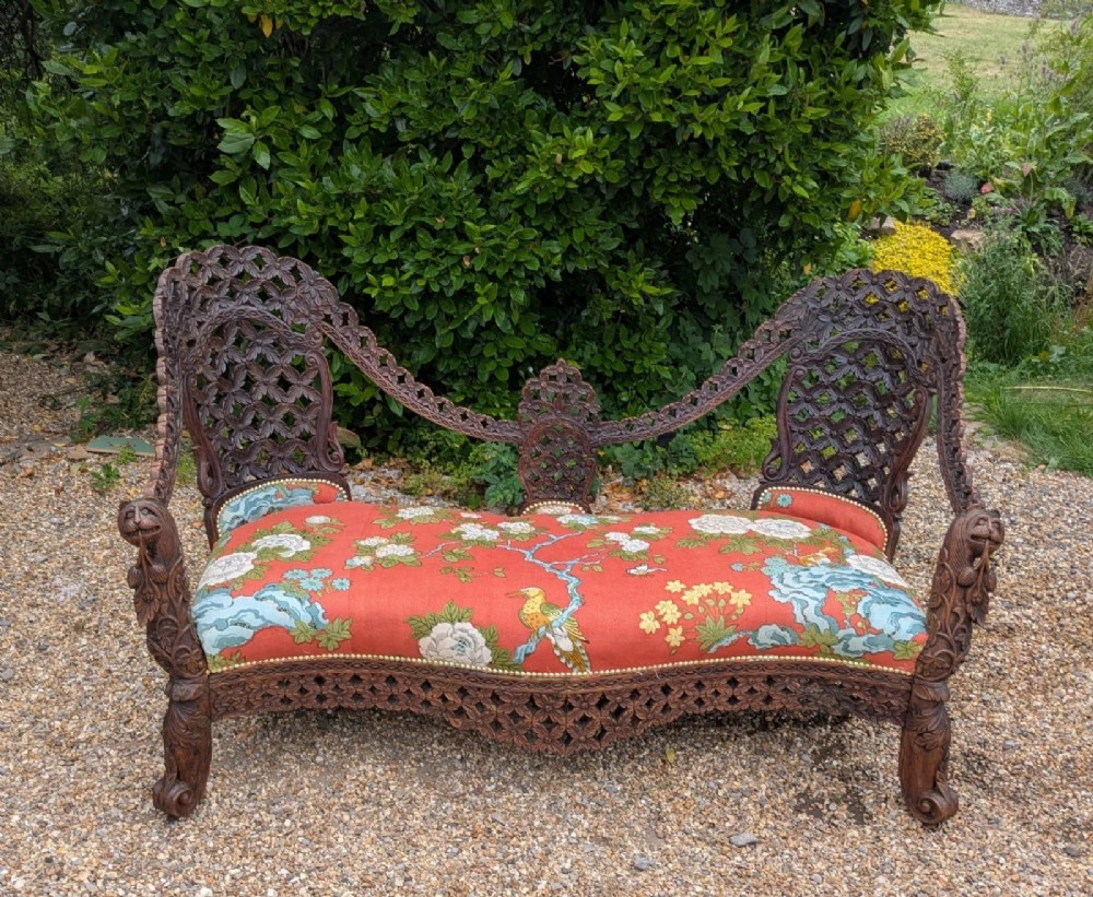 An antique Anglo-Indian carved hardwood sofa featuring an ornate openwork pierced backrest, carved lion armrests, and a patterned coral upholstered seat. View V