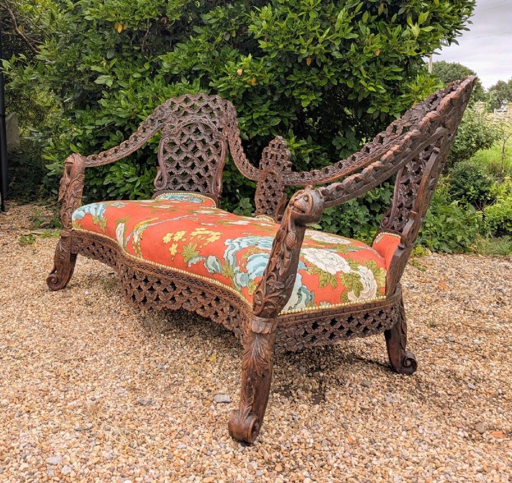 An antique Anglo-Indian carved hardwood sofa featuring an ornate openwork pierced backrest, carved lion armrests, and a patterned coral upholstered seat. View VII
