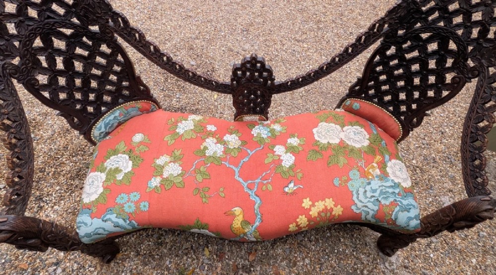 An antique Anglo-Indian carved hardwood sofa featuring an ornate openwork pierced backrest, carved lion armrests, and a patterned coral upholstered seat. View IX