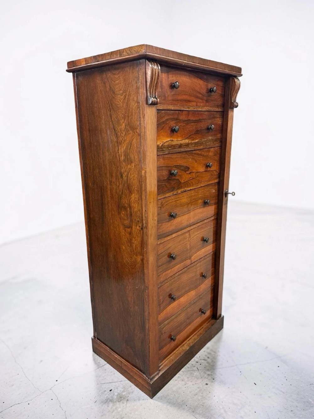 An antique Victorian rosewood Wellington chest featuring seven stacked drawers, turned wooden knobs, carved upper corbels, and a side locking bar.