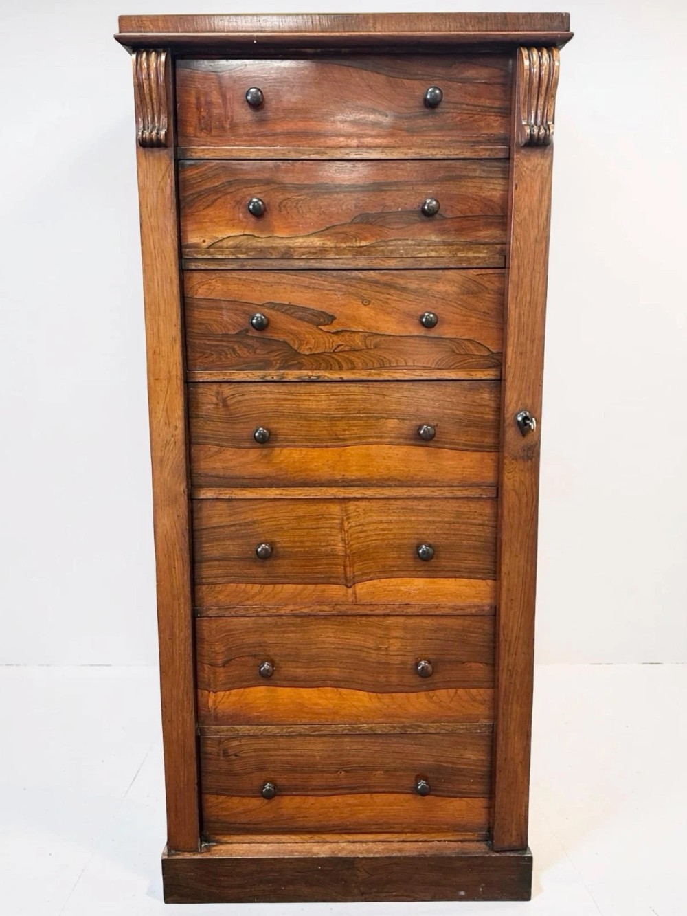 An antique Victorian rosewood Wellington chest featuring seven stacked drawers, turned wooden knobs, carved upper corbels, and a side locking bar. View II