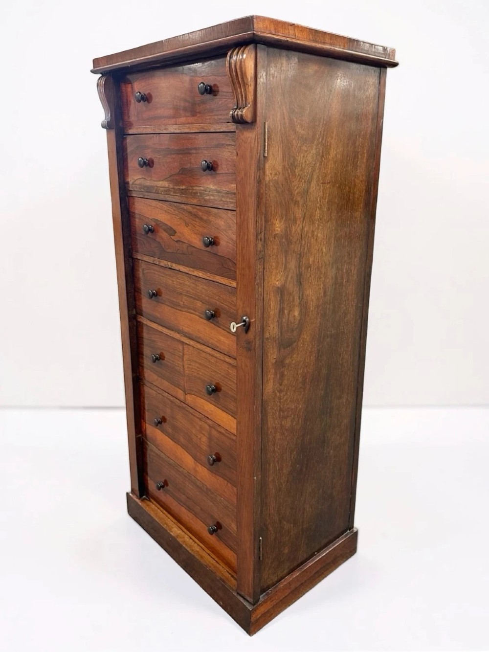An antique Victorian rosewood Wellington chest featuring seven stacked drawers, turned wooden knobs, carved upper corbels, and a side locking bar. View III