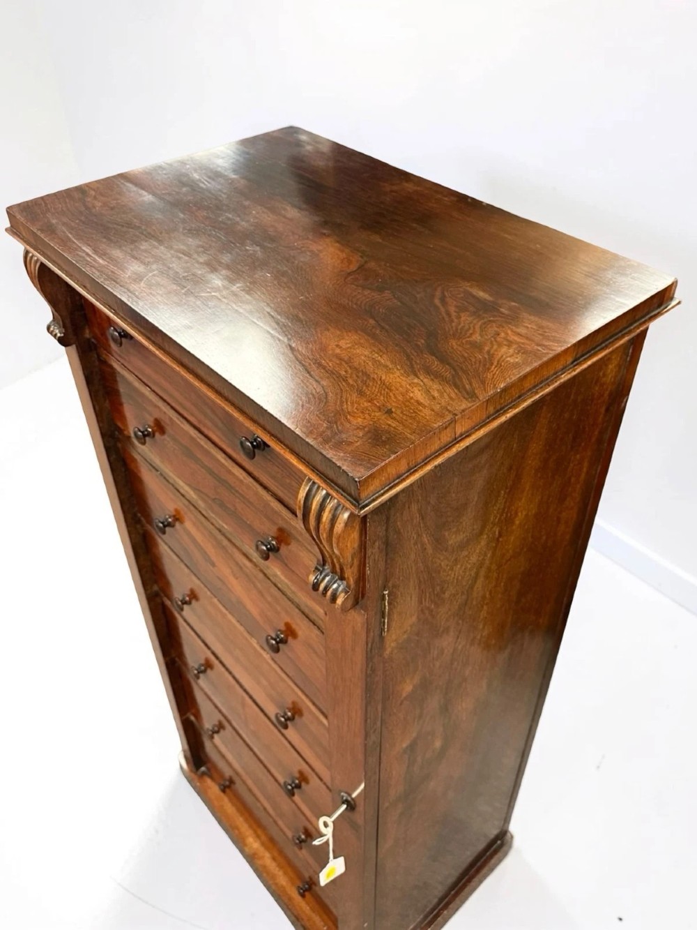 An antique Victorian rosewood Wellington chest featuring seven stacked drawers, turned wooden knobs, carved upper corbels, and a side locking bar. View X