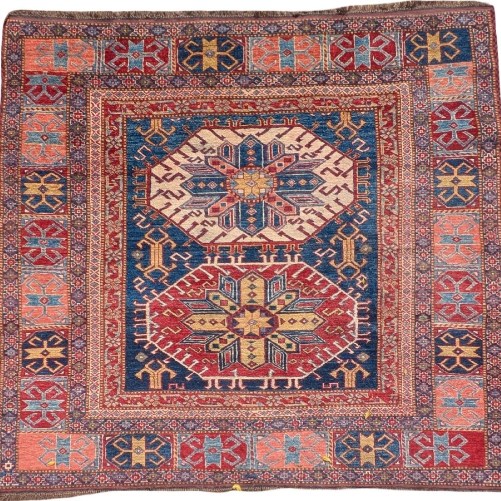 A square antique Caucasian Kazak wool rug featuring two prominent geometric star medallions on an indigo blue field surrounded by geometric borders in terracotta red, cream, and blue.