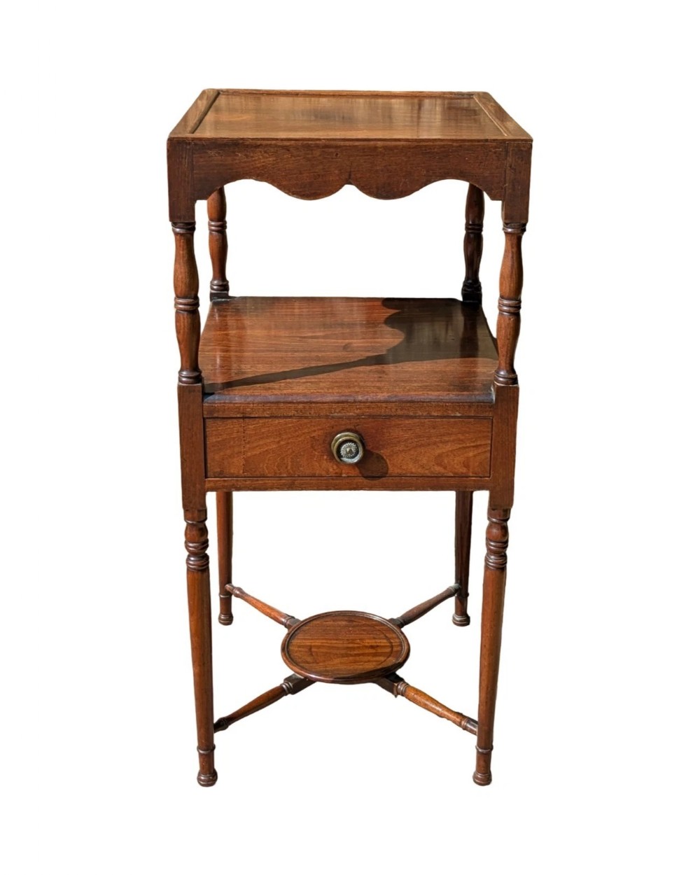 A George III mahogany bedside table with an upper tray top, a middle shelf, a single drawer with a central brass handle, and turned legs joined by an X-stretcher with a small circular tier.