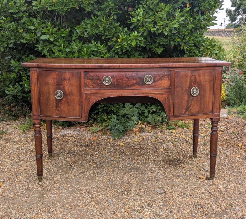 A Regency period mahogany sideboard featuring turned legs on brass castors, central drawer, flanking cupboard doors, and circular brass ring handles.