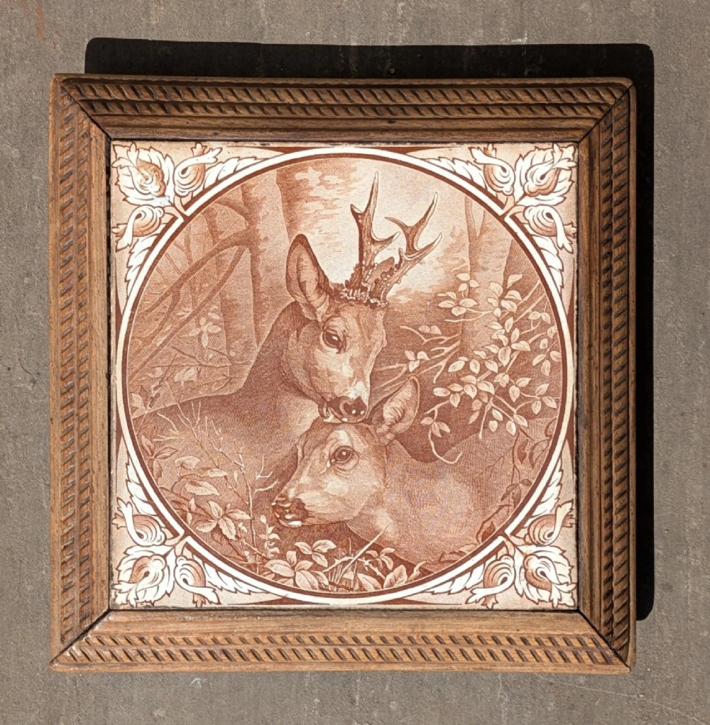 A square antique ceramic tile depicting a stag and doe in a woodland setting, framed within carved oak rope-twist moulding.
