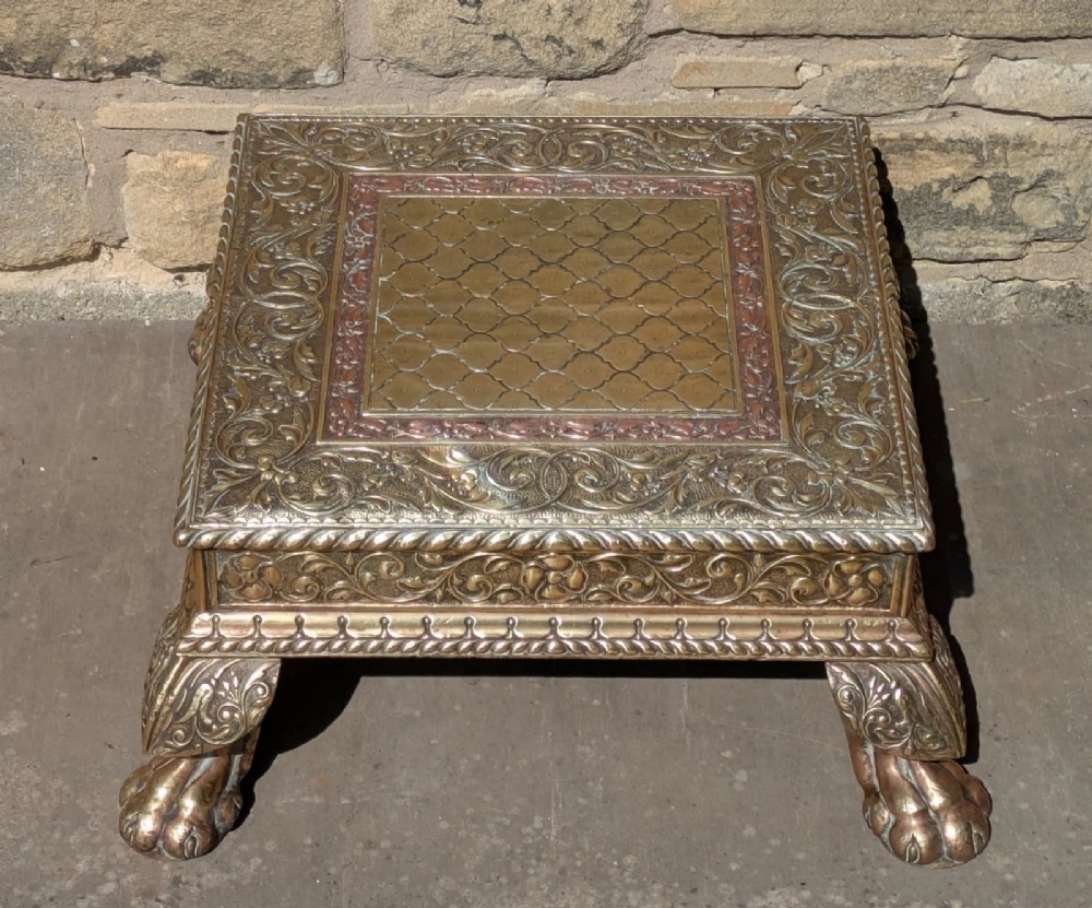 A square antique Indian low table with embossed brass and copper metal cladding, floral foliage motifs, a central geometric panel, and four claw feet. View III