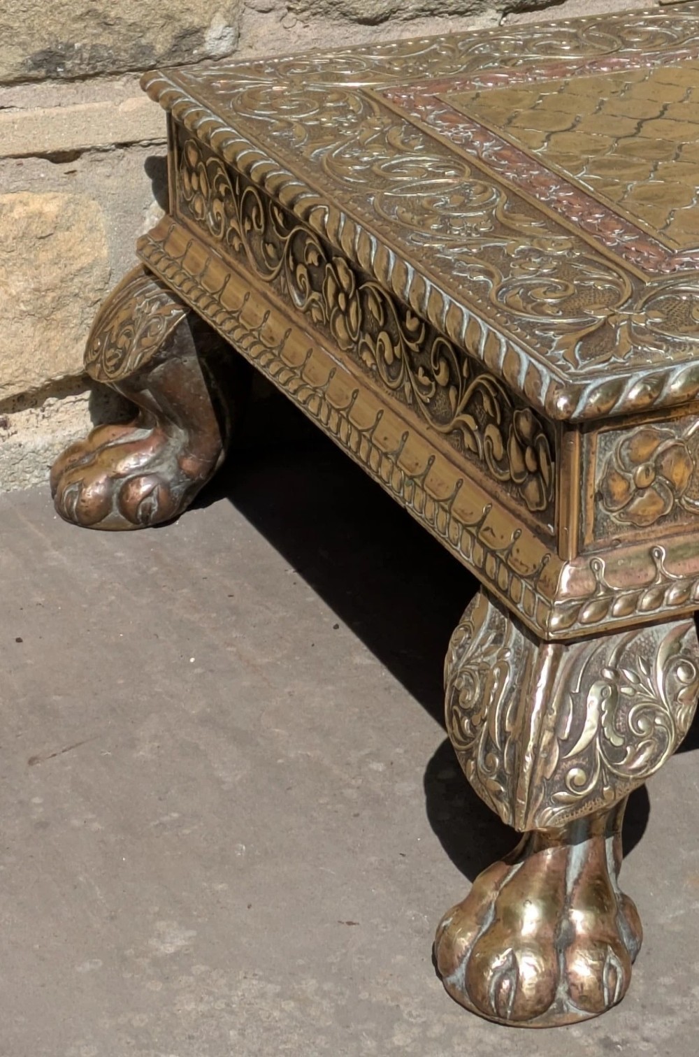 A square antique Indian low table with embossed brass and copper metal cladding, floral foliage motifs, a central geometric panel, and four claw feet. View IV