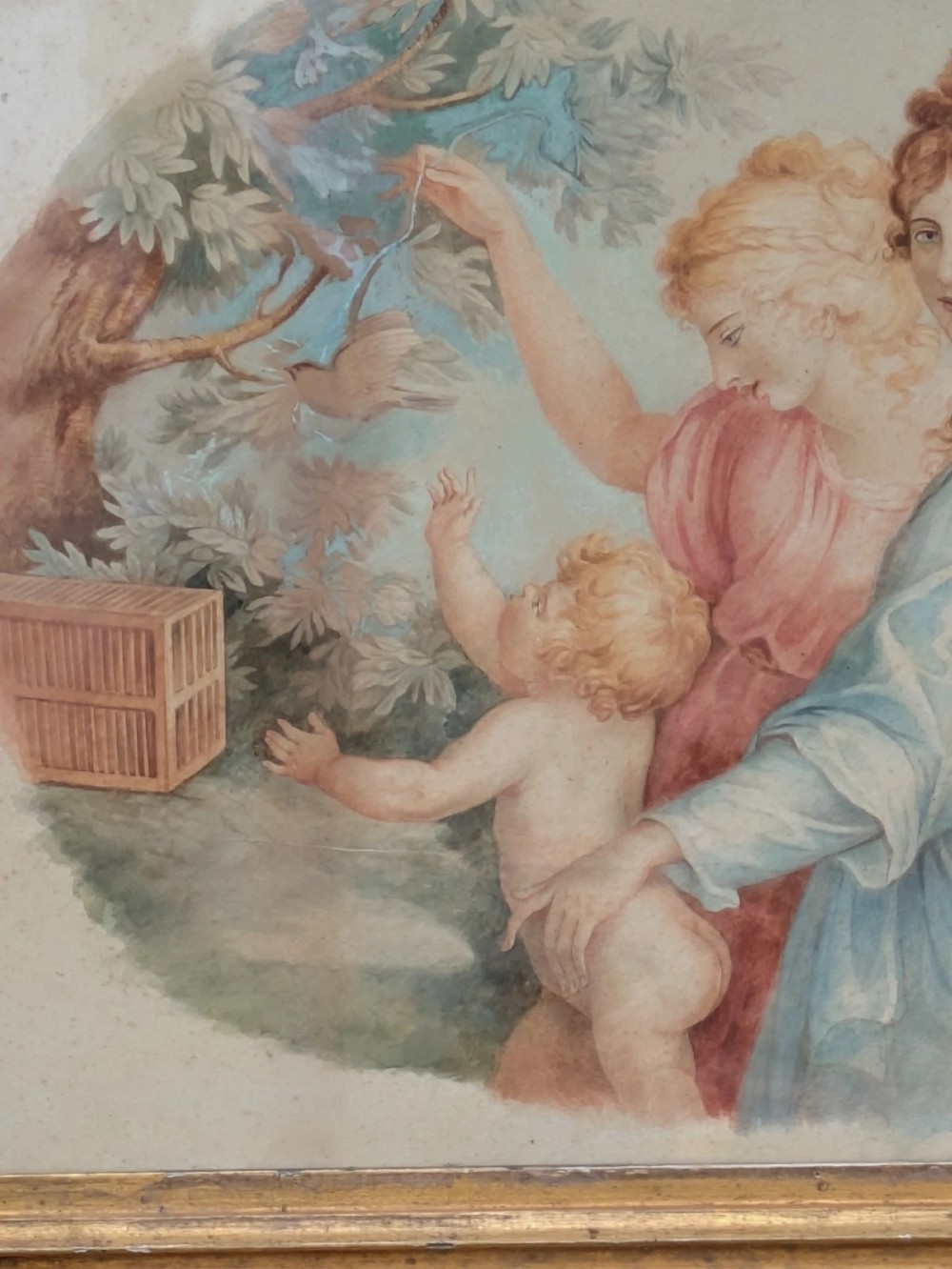 A framed classical watercolour painting depicting two draped female figures and a child beneath trees, presented in a moulded gilt frame. View V