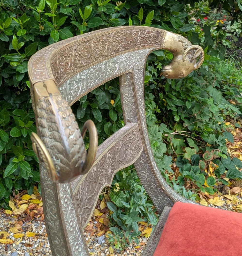 An Indian ceremonial chair wrapped in embossed silver metal with sculpted rams head terminals and a red velvet upholstered seat. View II