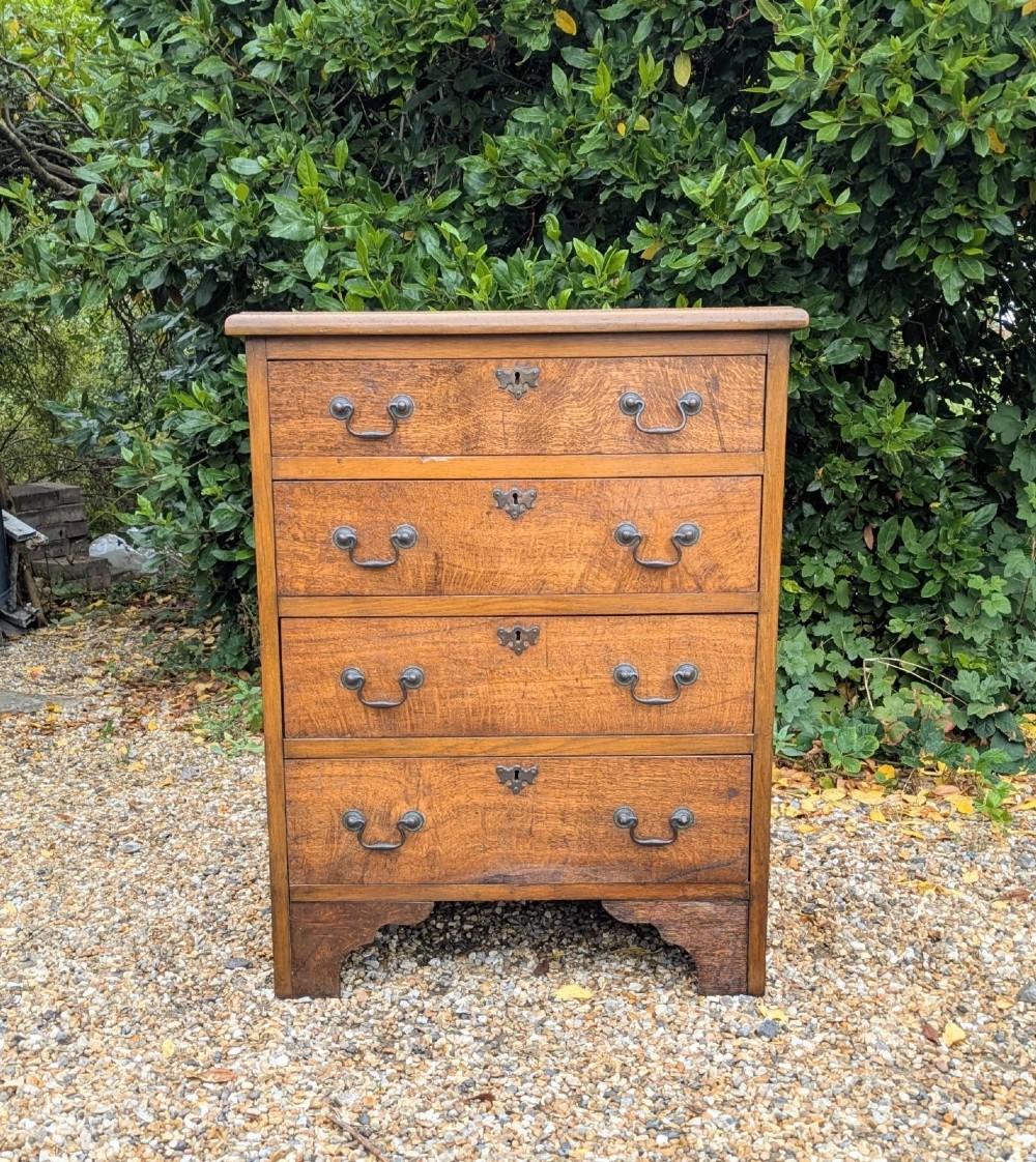 A compact oak bachelors chest of drawers featuring four graduated drawers with metal drop handles and keyhole escutcheons on a shaped bracket base. View VII