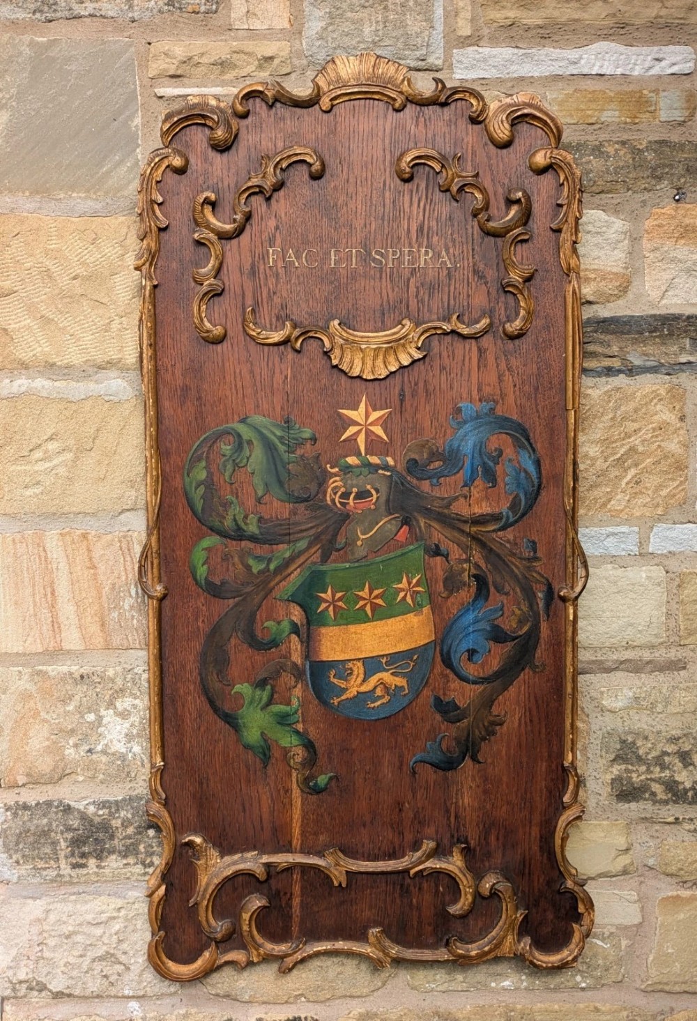 A rectangular carved oak wall plaque featuring a hand-painted heraldic coat of arms, gilt scroll moulding, and a Latin motto.
