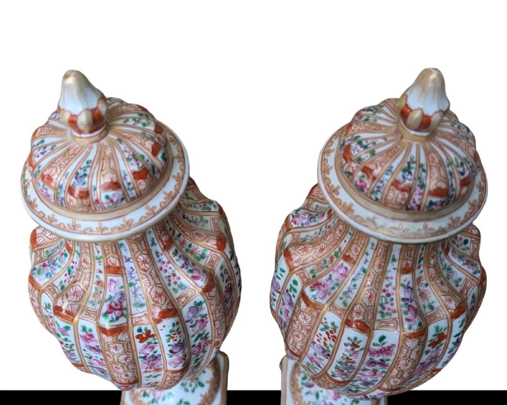 A pair of hand-painted porcelain lidded vases featuring fluted bodies, intricate floral motifs, geometric orange bands, and gilded details on square pedestals. View X