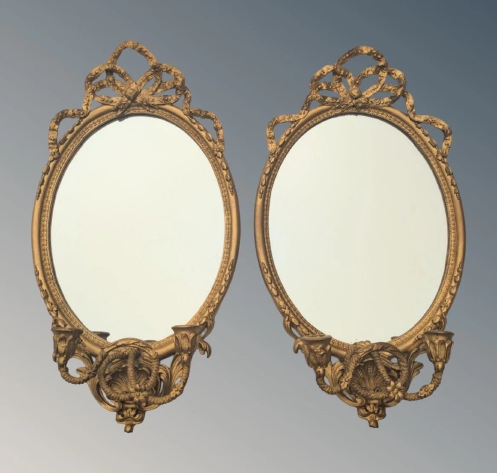A matching pair of oval giltwood girandole wall mirrors featuring ribbon cresting and twin candle sconces at the base.