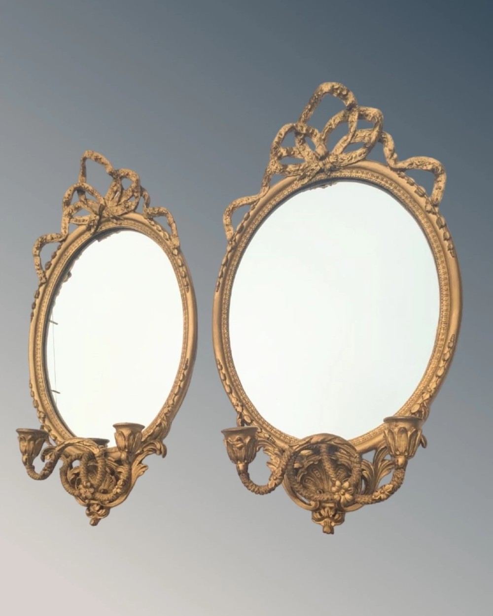 A matching pair of oval giltwood girandole wall mirrors featuring ribbon cresting and twin candle sconces at the base. View IV