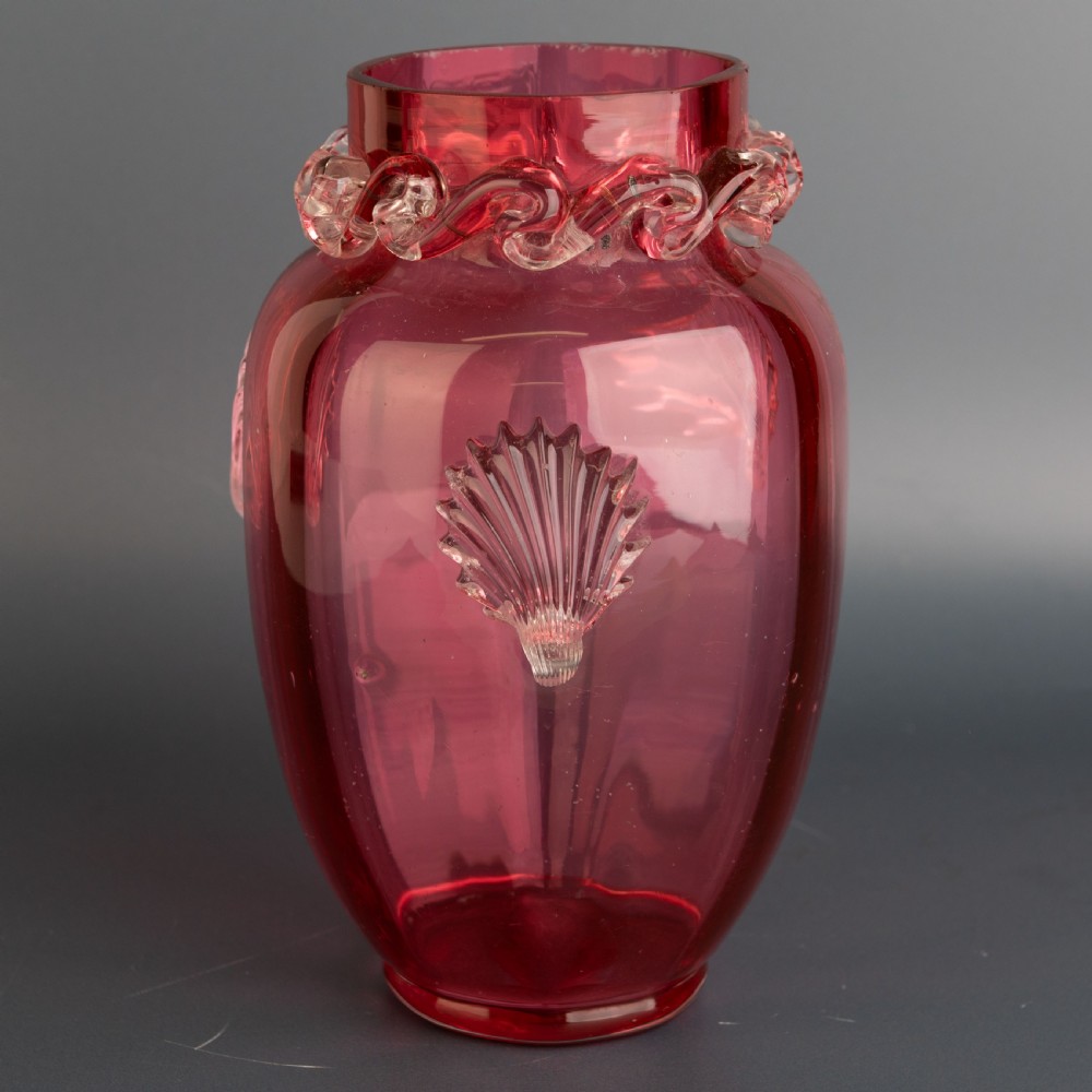 A Victorian hand-blown cranberry glass vase featuring an applied clear glass shell motif on the front and a crimped clear glass collar around the neck.