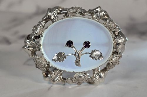 fig 1. An oval translucent chalcedony brooch set within a foliate silver frame, featuring a silver botanical spray with two purple amethyst stones at the centre.