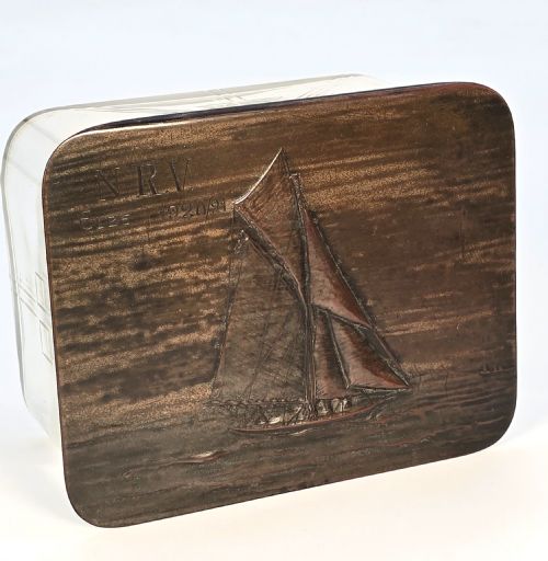 fig 1. A clear glass rectangular box with a patinated bronze lid featuring a raised relief depiction of a sailing boat.