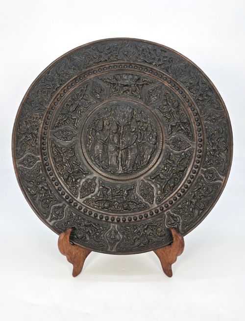 fig 1. A circular dark metal charger featuring intricate repousse depicting central figures under a tree surrounded by concentric decorative borders.