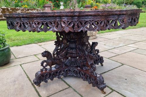 fig 1. A 19th-century Anglo-Indian carved padauk wood centre table featuring a scalloped top, deeply carved pierced apron of foliage, and a pedestal base supported by carved mythical lion feet with brass castors.