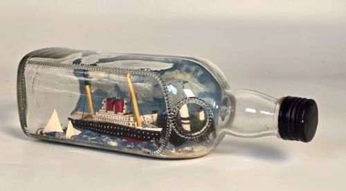 fig 1. A horizontal clear glass bottle containing a detailed 1930s folk art diorama of a black, white, and red steamship on painted blue water with a lighthouse backdrop.