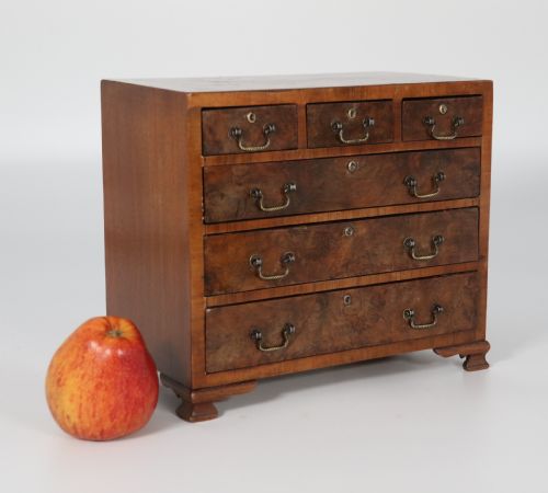 fig 1. A miniature George II style chest of drawers crafted from figured walnut with brass drop handles, standing 20cm high beside a red apple for scale.