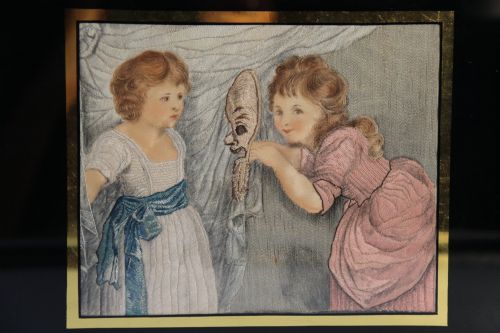 fig 1. A framed Georgian silk needlework picture depicting two young children with a mask, mounted in a black eglomise frame with gold leaf detailing.