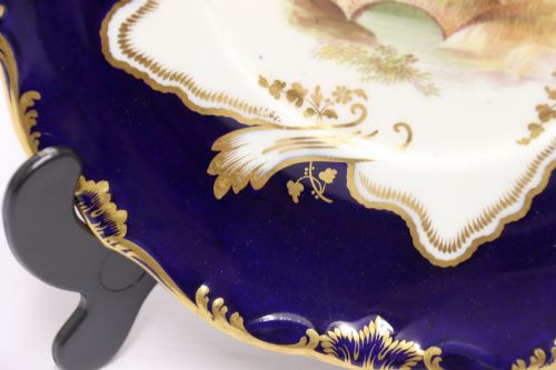 fig 1. A 19th-century Mintons porcelain dessert cream tureen with lid and handles positioned alongside its matching stand, decorated with cobalt blue ground, gold gilding, and hand-painted landscape scenes.