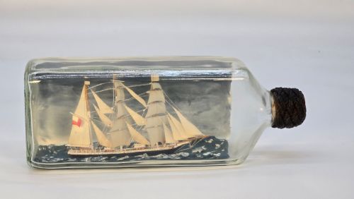 fig 1. A horizontal clear glass bottle containing a detailed miniature three-masted sailing ship diorama set upon painted blue ocean waves with a rope-bound neck stopper.