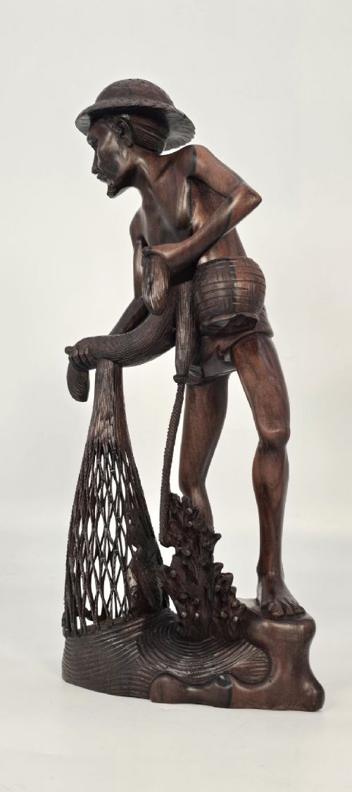 fig 1. A hand-carved dark hardwood sculpture depicting a standing fisherman holding a woven net and basket, set on an organic carved base.
