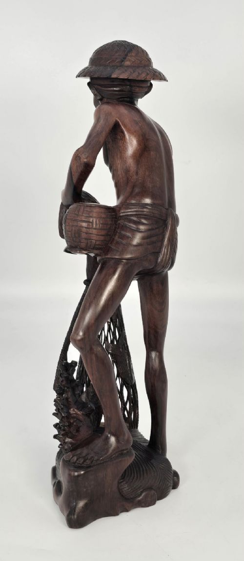 A hand-carved dark hardwood sculpture depicting a standing fisherman holding a woven net and basket, set on an organic carved base. View XI