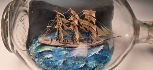 fig 2. A vintage clear glass bottle containing a detailed miniature three-masted sailing vessel set upon painted blue waves against a dark background.