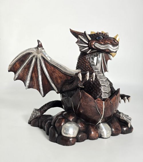 fig 1. A carved dark hardwood sculpture depicting a dragon hatching from an egg, detailed with silvered and brass metal mounts on its wings, underbelly, and claws.