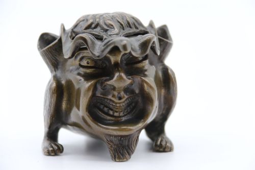 fig 1. A dark cast bronze novelty inkwell sculpted in the form of a grotesque grinning face with textured hair and small footed base.