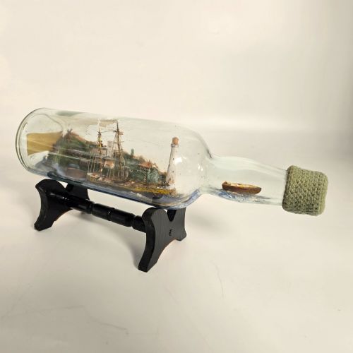 fig 1. A clear glass bottle containing a handcrafted folk art diorama of a sailing vessel, lighthouse, and coastal town, supported by a black wooden display stand.