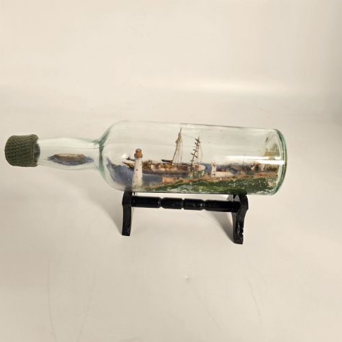 fig 2. A clear glass bottle containing a handcrafted folk art diorama of a sailing vessel, lighthouse, and coastal town, supported by a black wooden display stand.