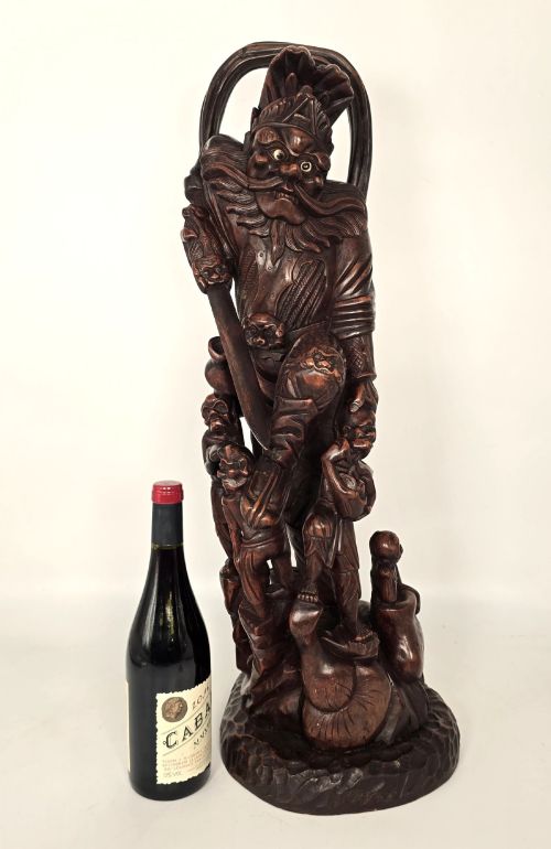 fig 1. A tall carved dark hardwood Chinese sculpture depicting a Guardian King wearing detailed armour and holding a sword over attendant figures.