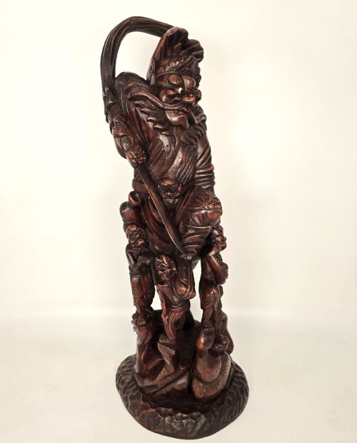 fig 2. A tall carved dark hardwood Chinese sculpture depicting a Guardian King wearing detailed armour and holding a sword over attendant figures.