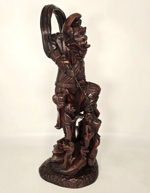 fig 3. A tall carved dark hardwood Chinese sculpture depicting a Guardian King wearing detailed armour and holding a sword over attendant figures.