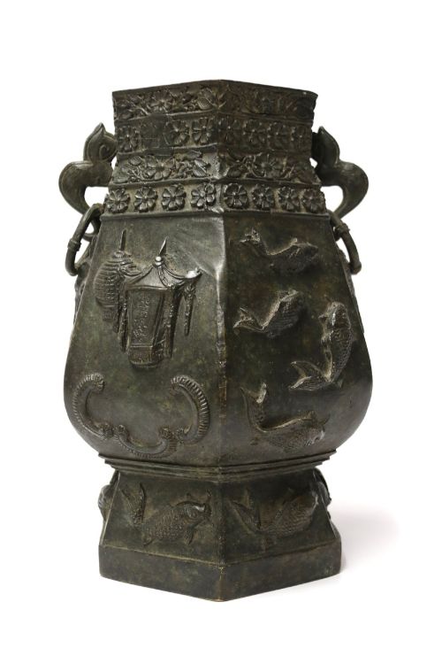 fig 1. An antique hexagonal solid bronze vessel featuring relief carvings of fish, architectural structures, floral bands, and twin animal-head ring handles.