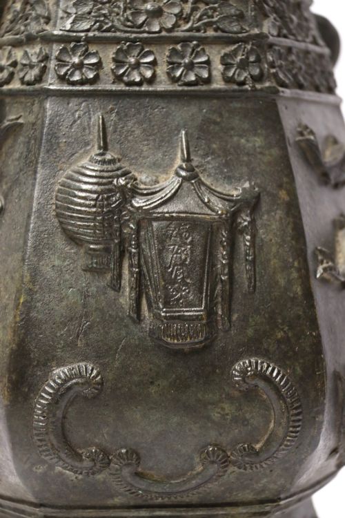 fig 2. An antique hexagonal solid bronze vessel featuring relief carvings of fish, architectural structures, floral bands, and twin animal-head ring handles.