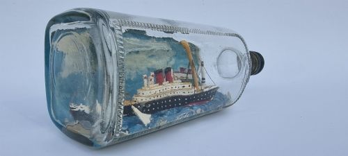 fig 1. A horizontal glass bottle containing a handcrafted 1930s steamship diorama with red funnels, a painted blue sea, and a lighthouse background.